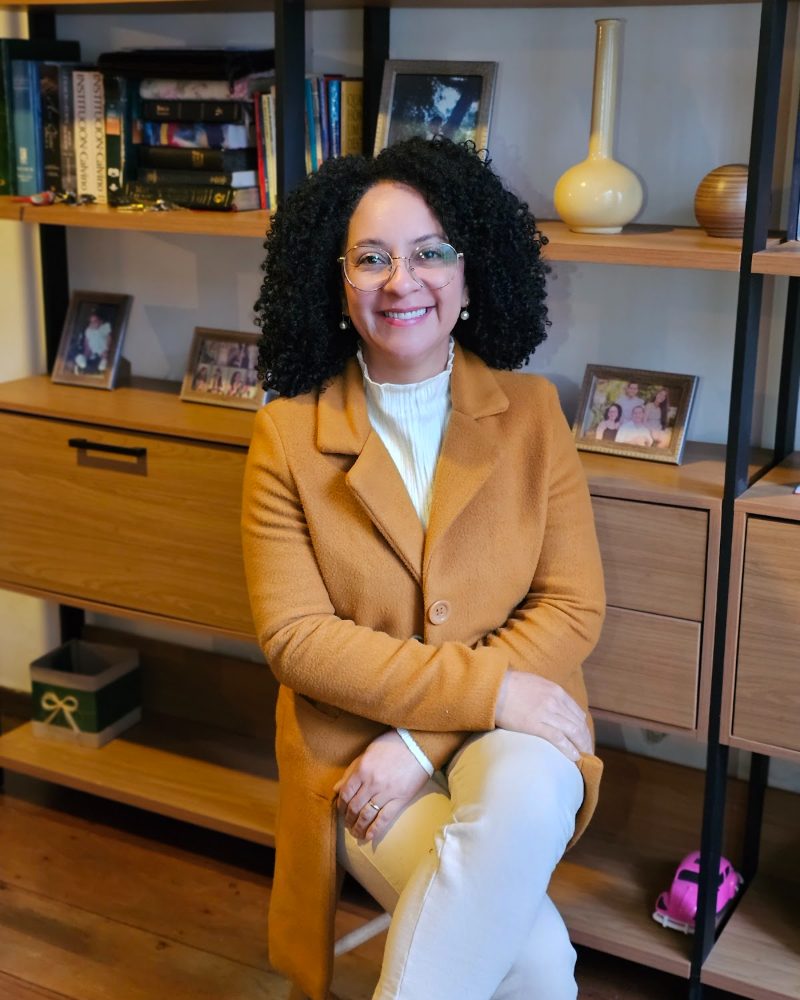 Suely Gusmão, psicóloga, sentada diante da estante do consultório, de casaco caramelo, sorrindo